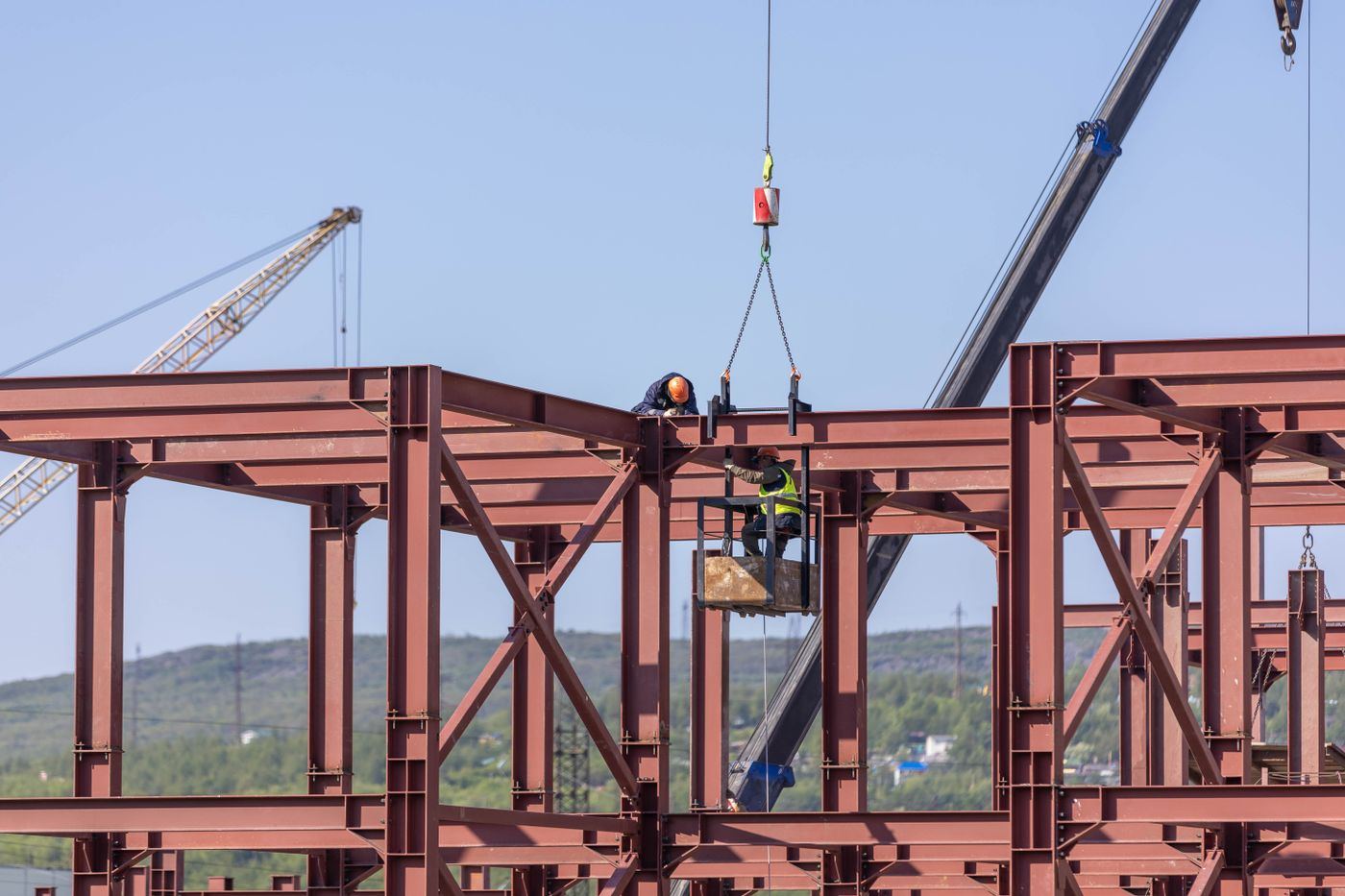 Two riggers landing a steel beam on a primed structural frame with a crane hook and chains overhead